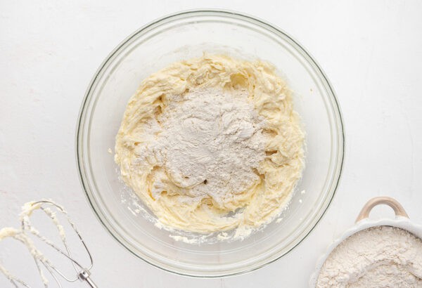 Flour added to the wet ingredients for spritz cookies in a glass mixing bowl.