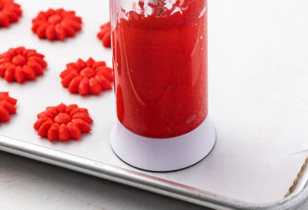 A spritz cookie press pressing out red flower shaped cooking on a parchment lined baking sheet.