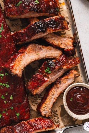 Overhead view of baked baby back ribs on a baking sheet with a side of BBQ sauce