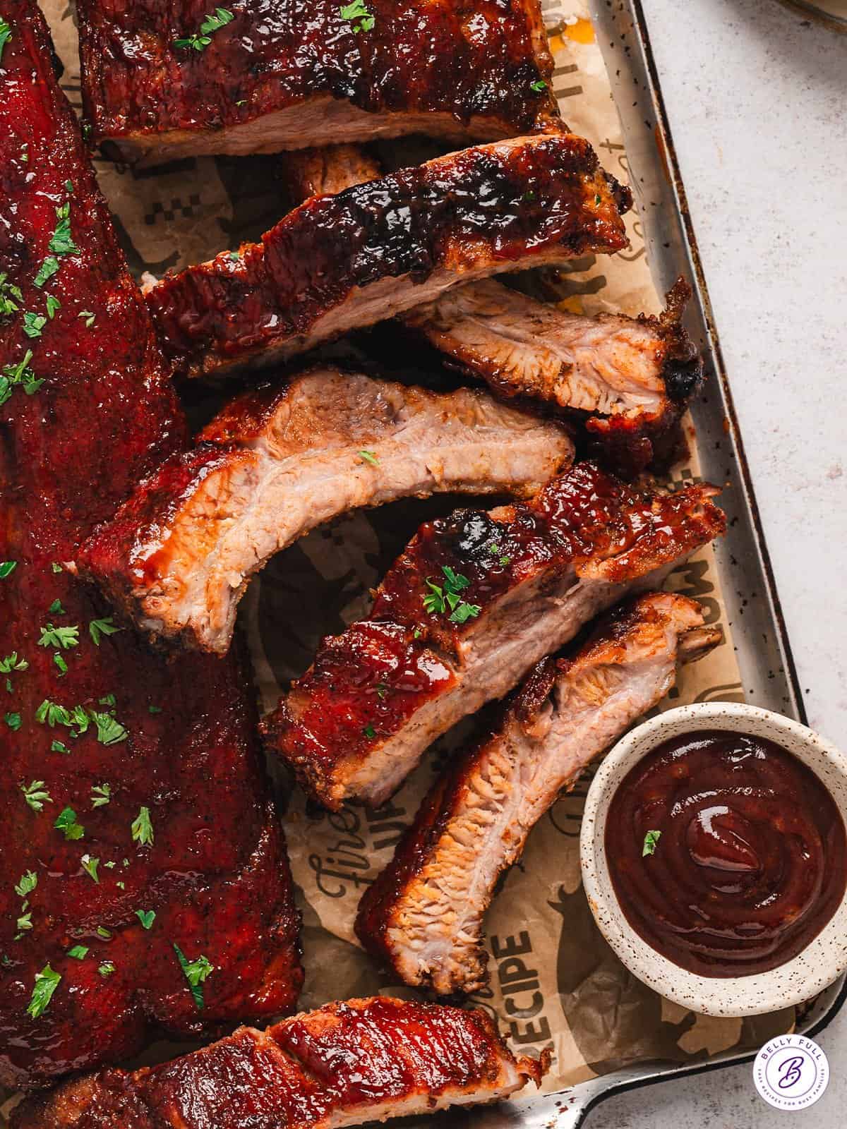 Overhead view of baked baby back ribs on a baking sheet with a side of BBQ sauce