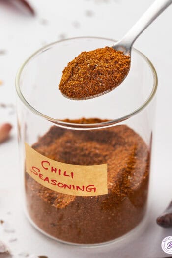 A glass jar of chili seasoning with a spoon of seasoning over the jar