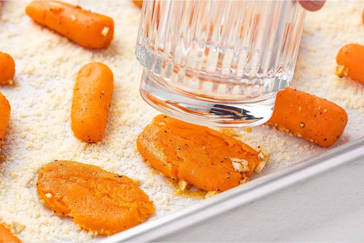 A glass smashing carrots on a baking sheet