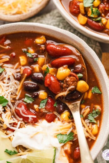 A spoon in a bowl of 7 can soup with shredded cheese, sour cream, and avocado slices