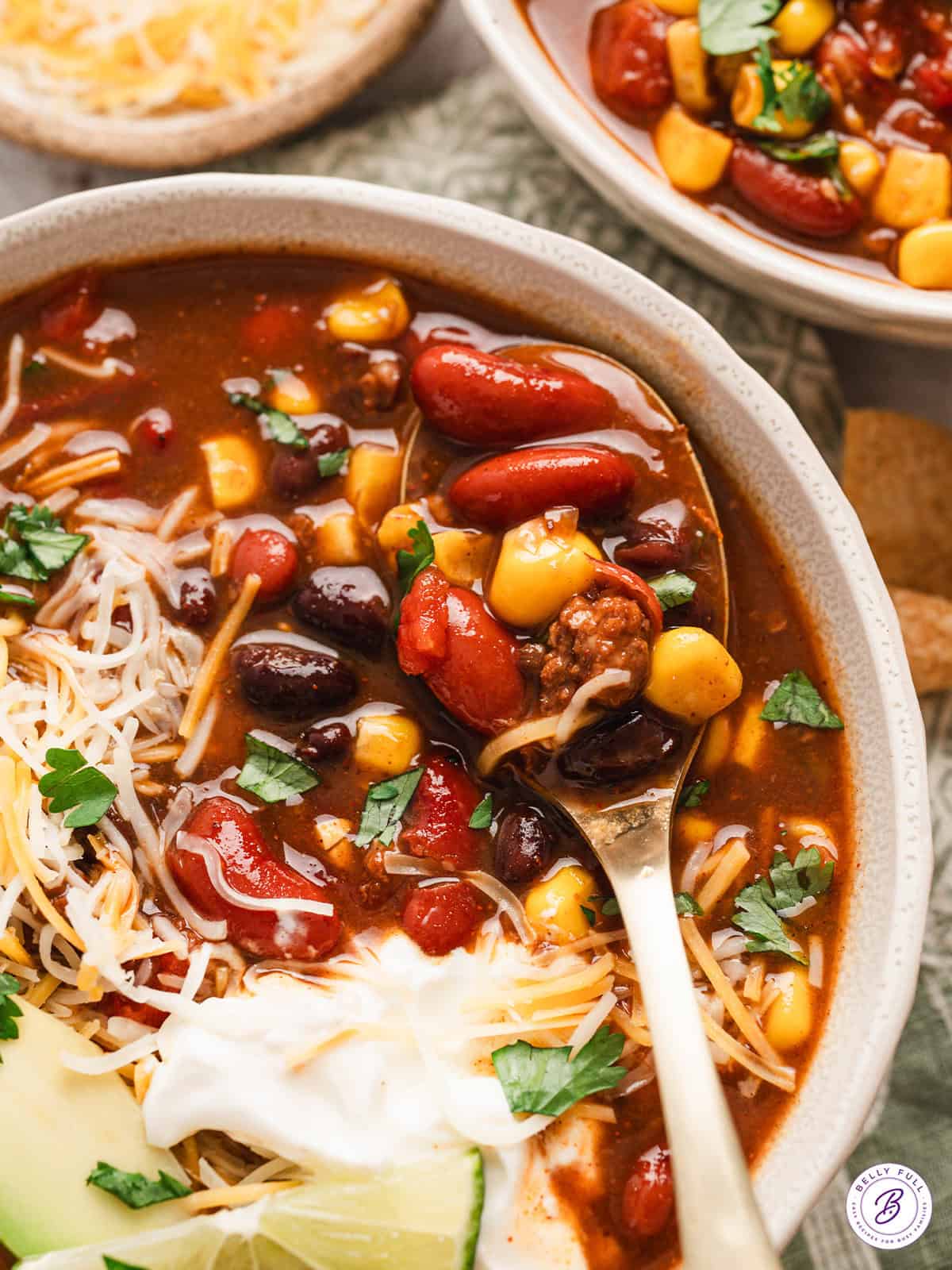 A spoon in a bowl of 7 can soup with shredded cheese, sour cream, and avocado slices