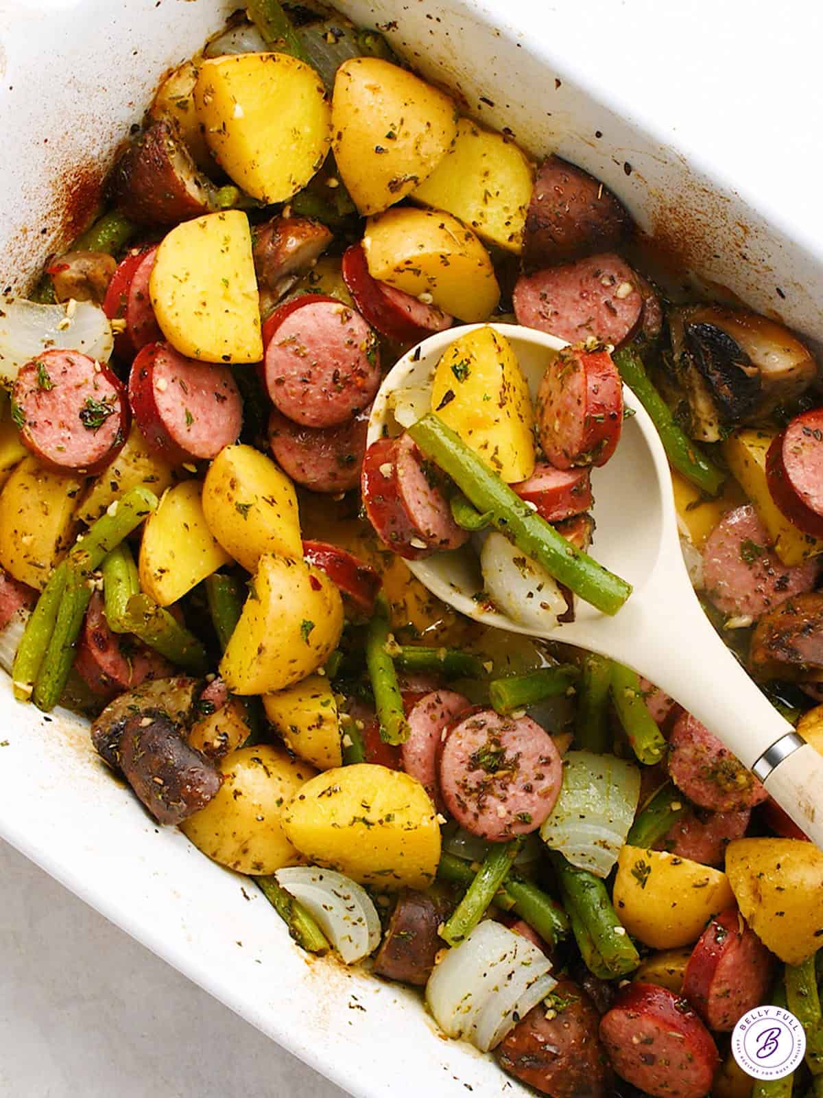 overhead baked sliced kielbasa and vegetables in casserole dish and serving spoon