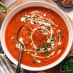 A bowl of roasted red pepper soup, topped with cream and fresh parsley, with a spoon