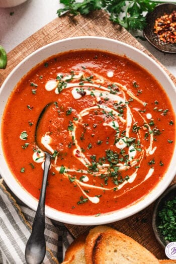 A bowl of roasted red pepper soup, topped with cream and fresh parsley, with a spoon