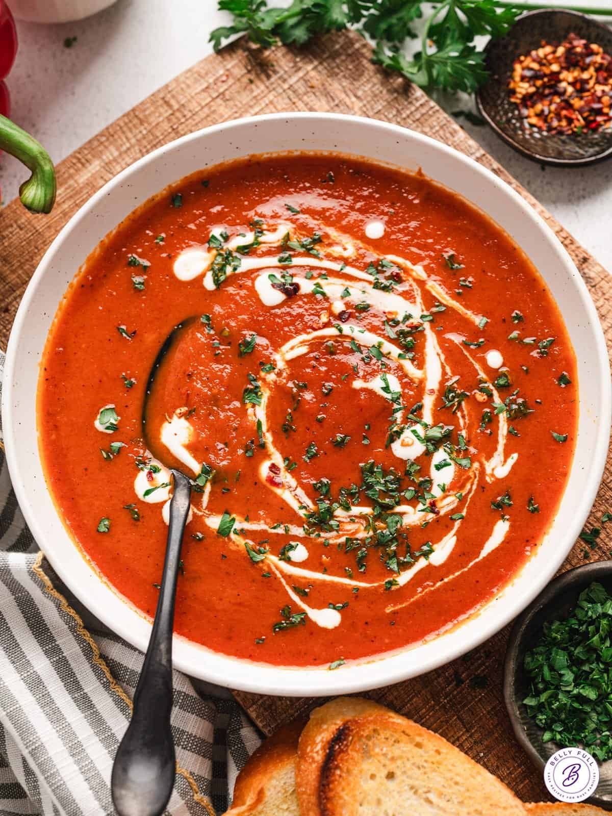 Overhead view of a bowl of roasted red pepper soup, topped with cream and fresh parsley, with a spoon.