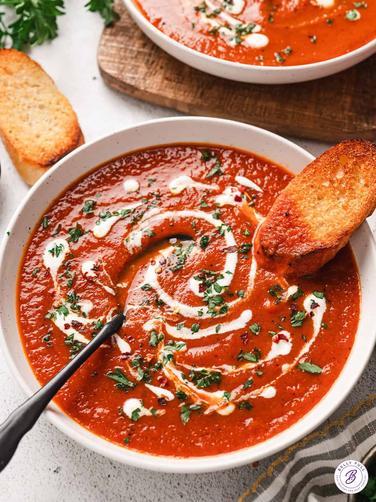 A bowl of roasted red pepper soup, topped with cream and fresh parsley, with a spoon and slice of bread.