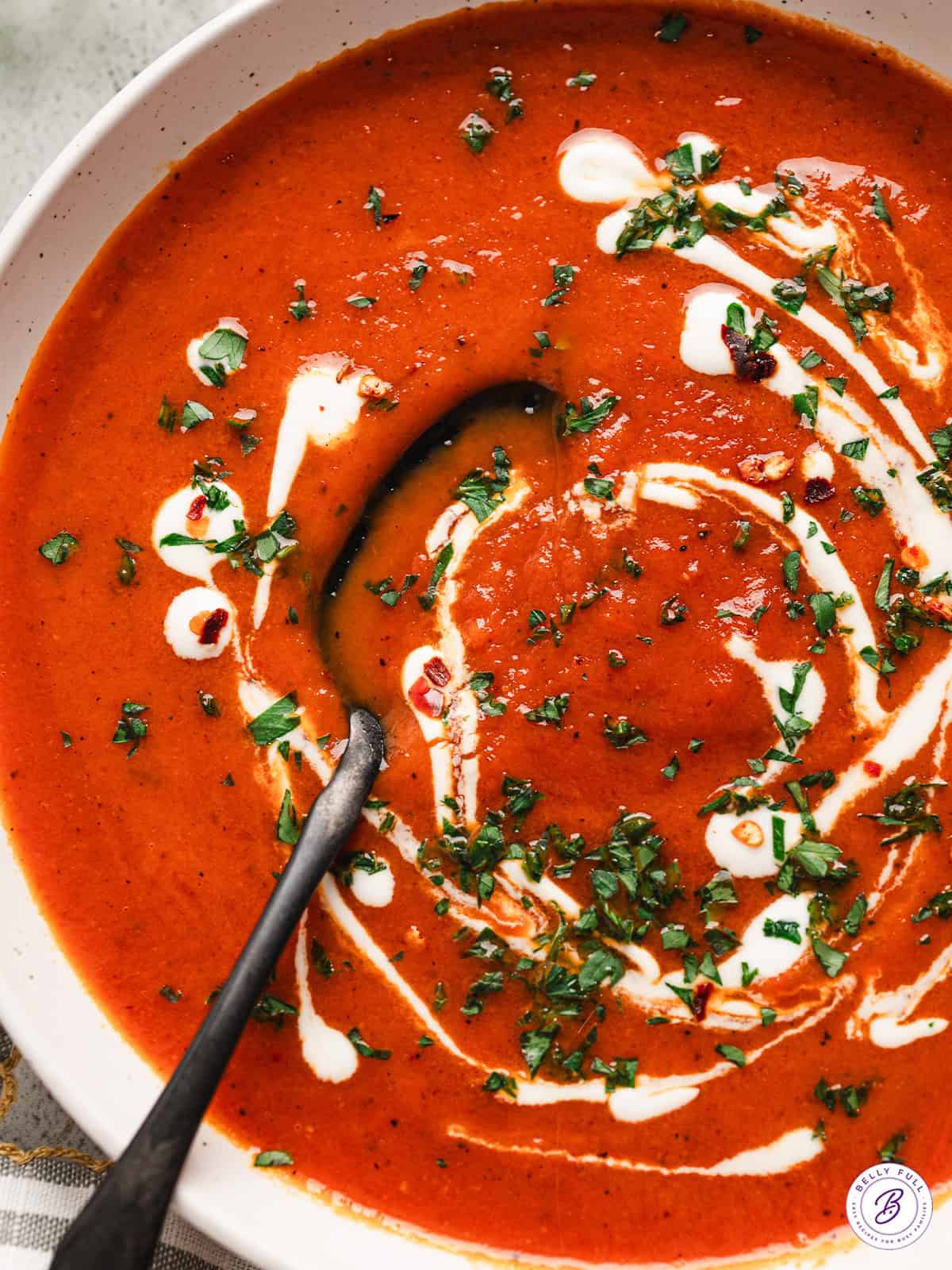 Overhead close up of a bowl of roasted red pepper soup, topped with cream and fresh parsley, with a spoon.