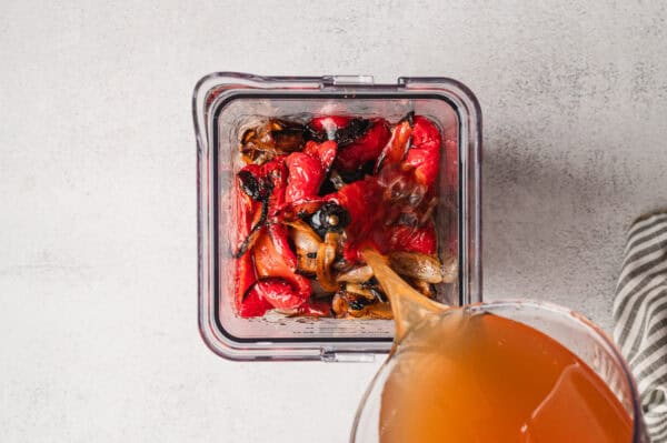 Vegetable broth being poured into a blender with roasted red pepper and sweet onion.