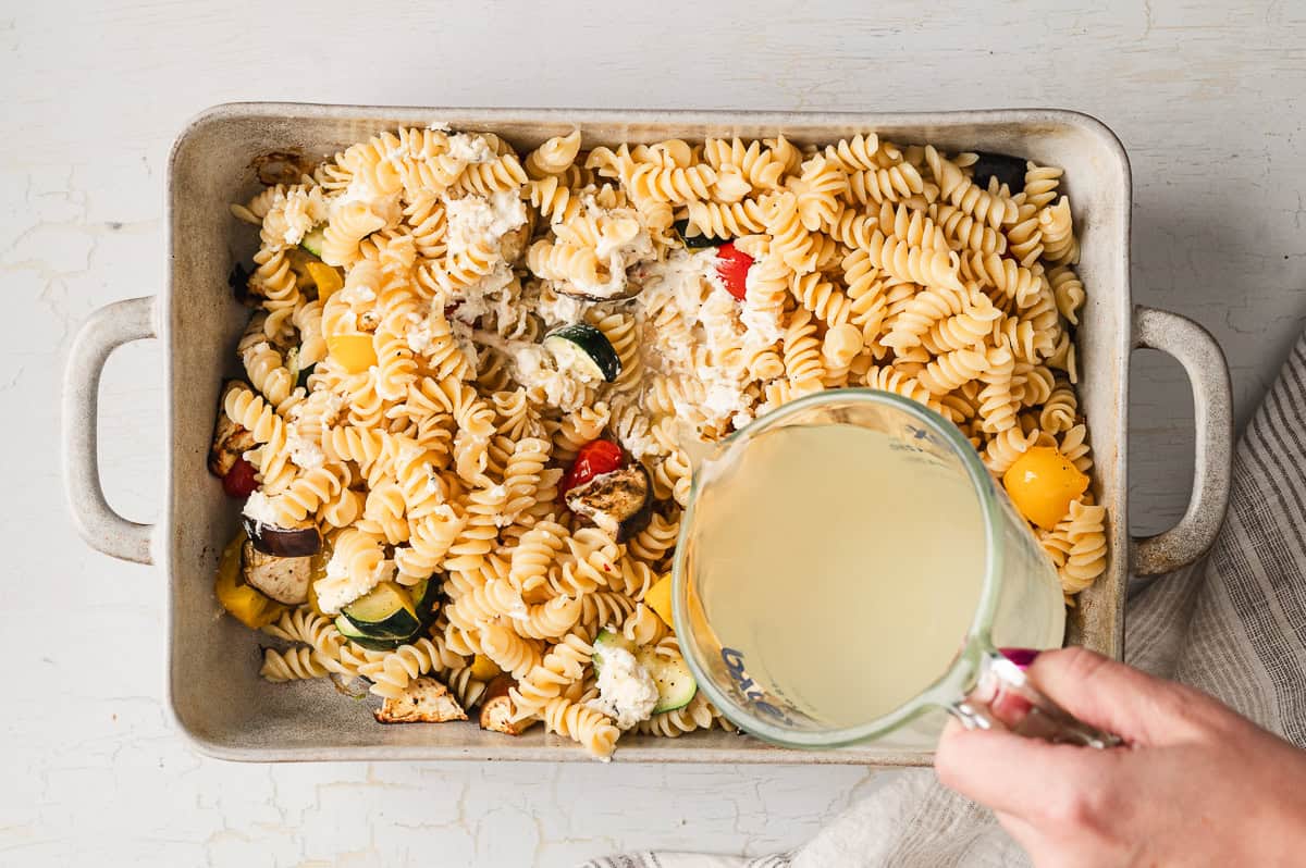 Pouring pasta water over penne and veggies on a baking sheet