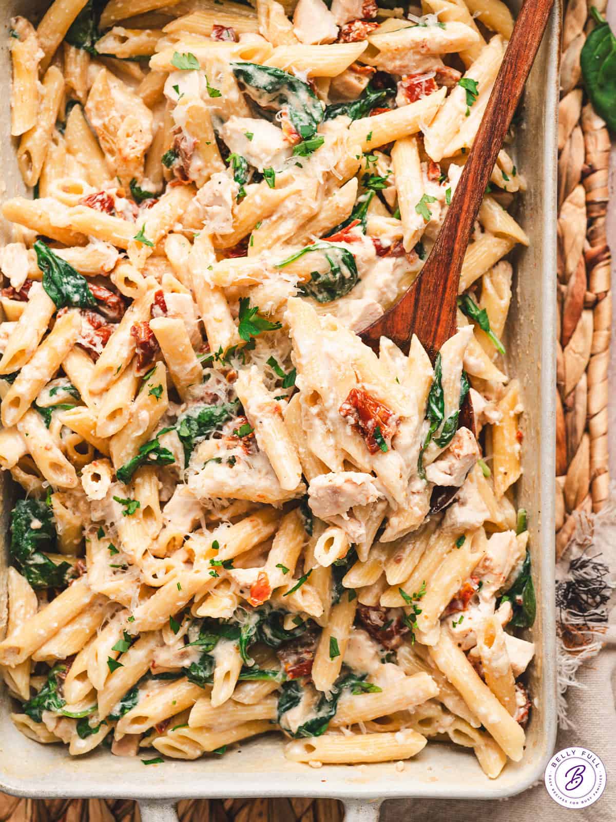 With penne pasta, spinach, and chicken tossed in a creamy sundried tomato sauce, this Baked Tuscan Chicken Pasta is an easy weeknight dinner. A wooden spoon in a casserole dish of baked Tuscan chicken pasta