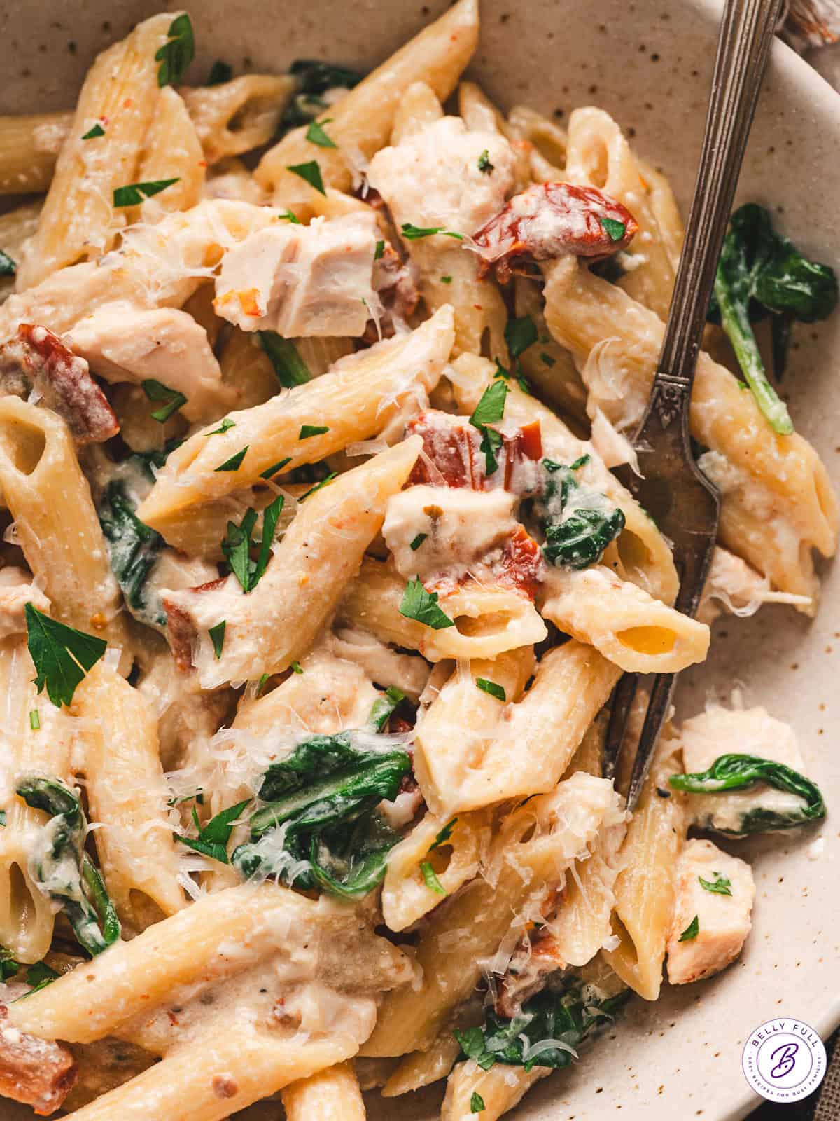 With penne pasta, spinach, and chicken tossed in a creamy sundried tomato sauce, this Baked Tuscan Chicken Pasta is an easy weeknight dinner. A wooden spoon stirring baked Tuscan chicken pasta