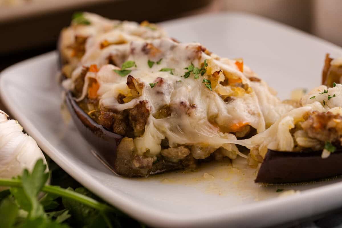 Close-up of cheesy stuffed eggplant topped with melted mozzarella and fresh parsley on a white plate, showing a hearty filling of ground meat and vegetables.