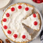 Overhead view of a Million Dollar Pie in a graham cracker crust topped with dollops of whipped cream and maraschino cherries, with one slice removed.