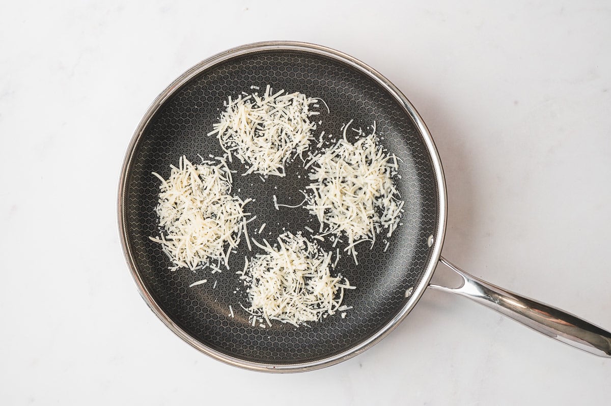 A nonstick skillet with piles of shredded cheese melting into round mounds to form frico nests.