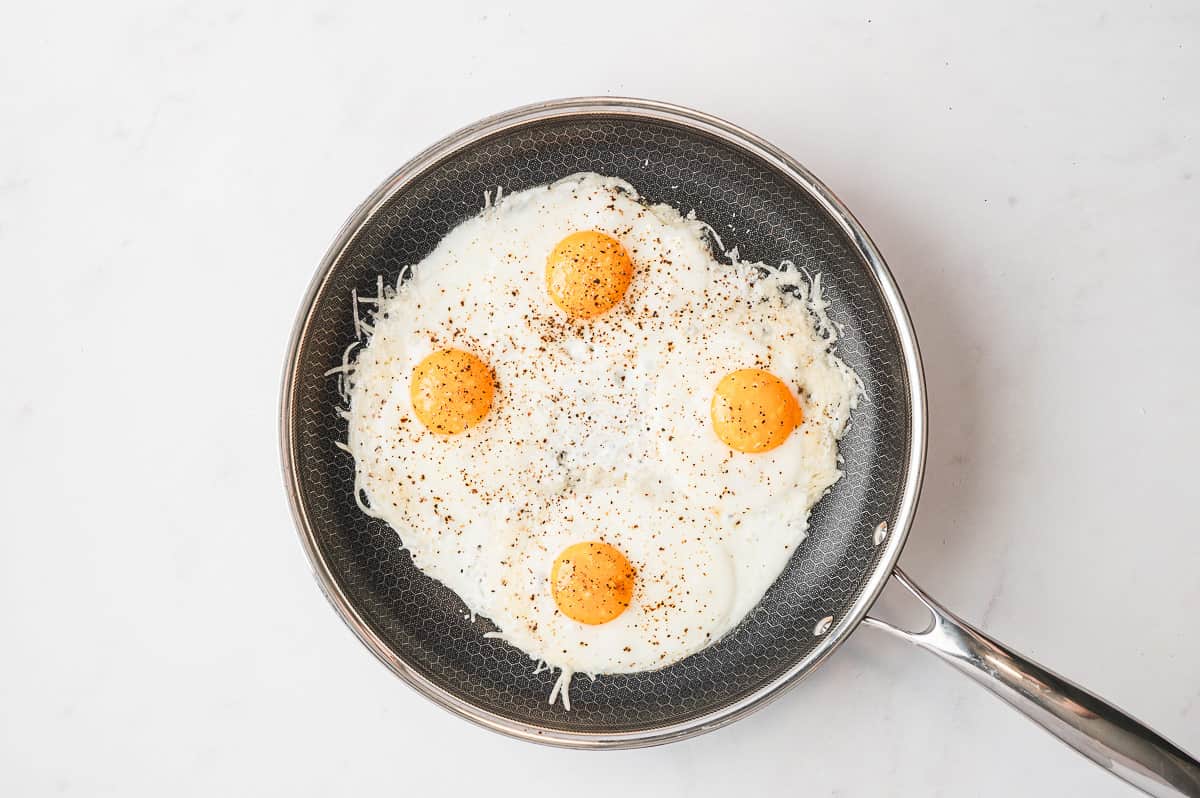 Eggs cracked into the center of each cheese nest in the skillet, cooking until set.