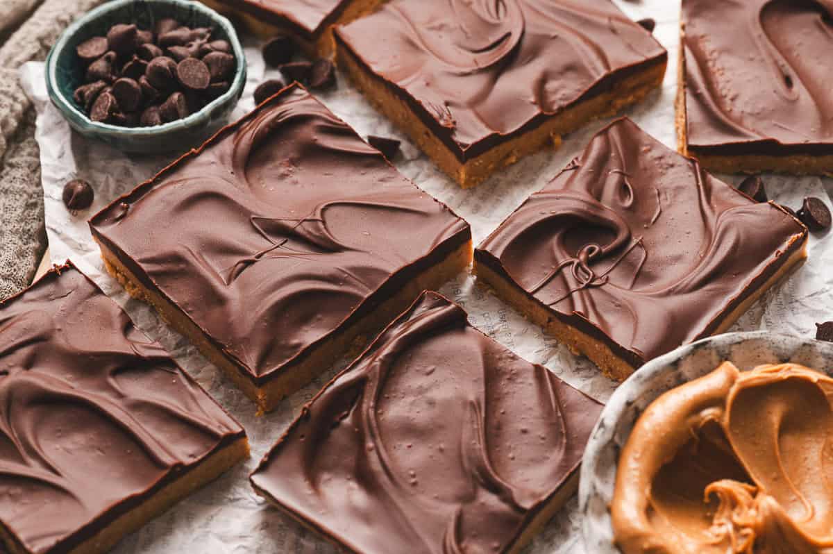 No-bake buckeye bars cut into squares with a smooth, glossy chocolate top and a thick peanut butter base, surrounded by scattered chocolate chips and a bowl of creamy peanut butter on parchment paper.