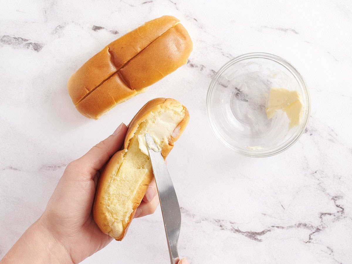 Hand spreading butter on the side of a split-top bun.