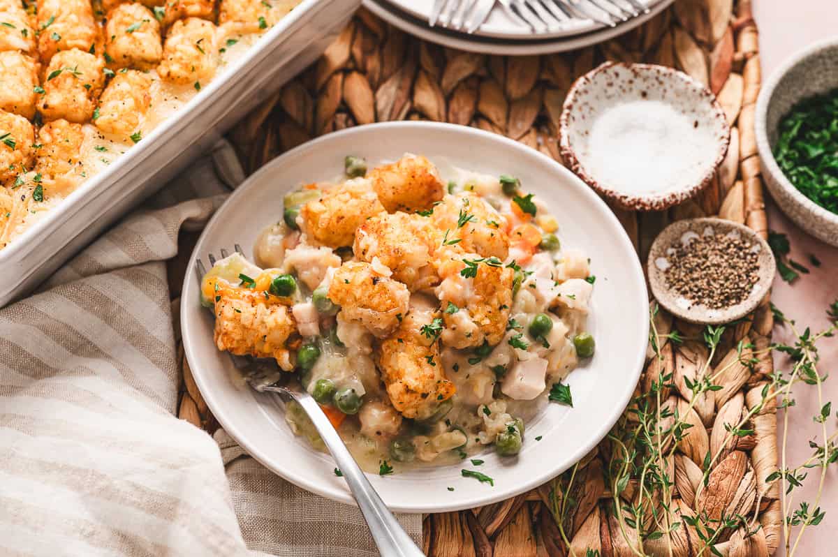 Close-up of a serving of chicken tot pie on a white plate, showing crispy golden tater tots on top of creamy chicken and vegetable filling with peas, carrots, and celery, garnished with fresh parsley.