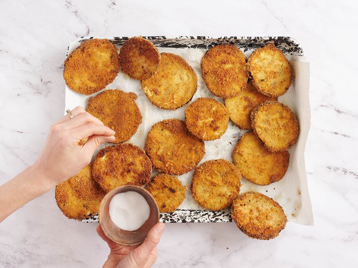 Hand sprinkling salt on freshly fried eggplant rounds.