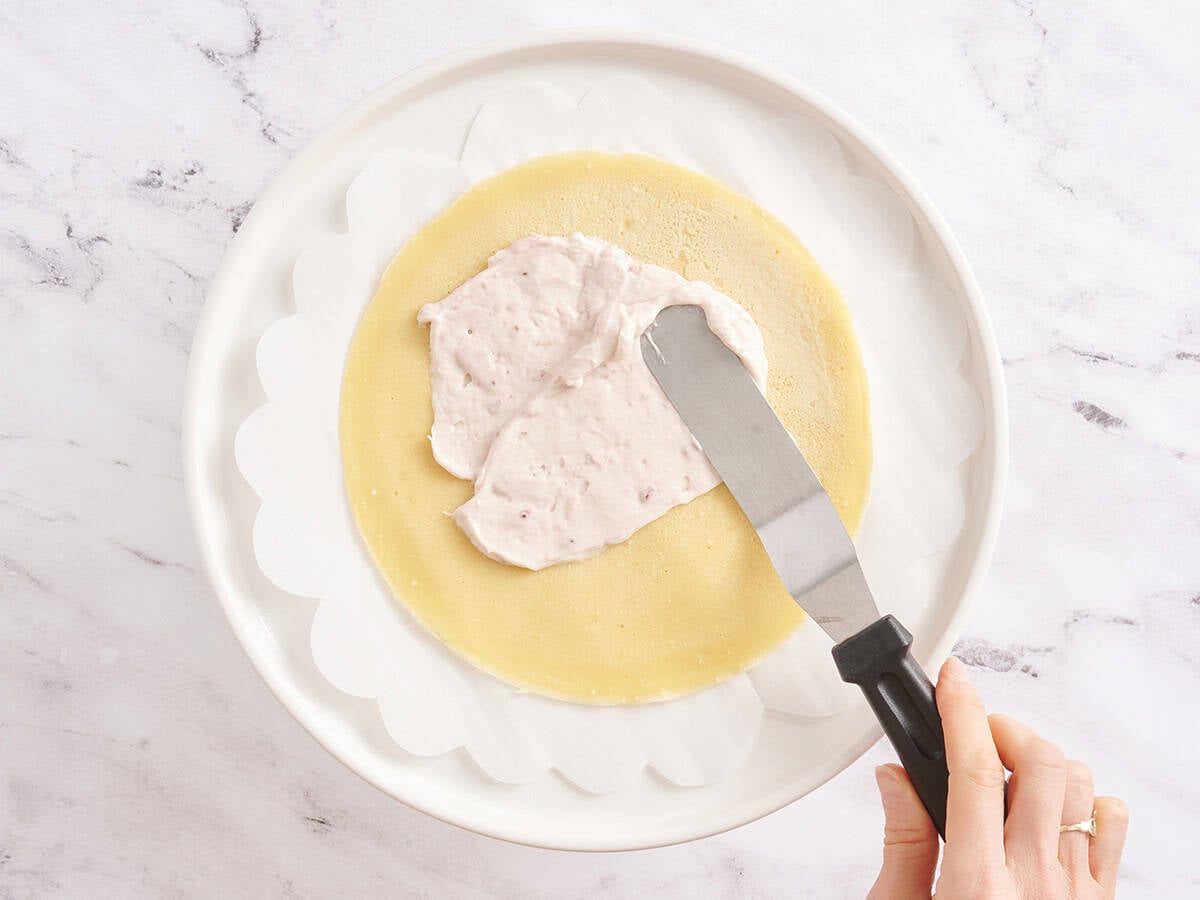 Spreading raspberry cream filling over a single crepe layer with an offset spatula on a parchment-lined plate.