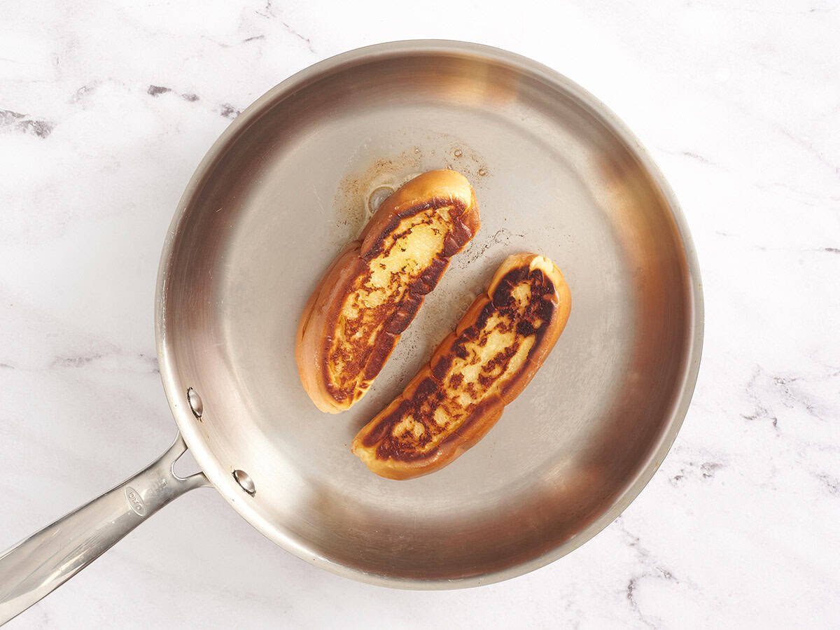 Buttered buns being toasted golden brown in a stainless steel skillet.