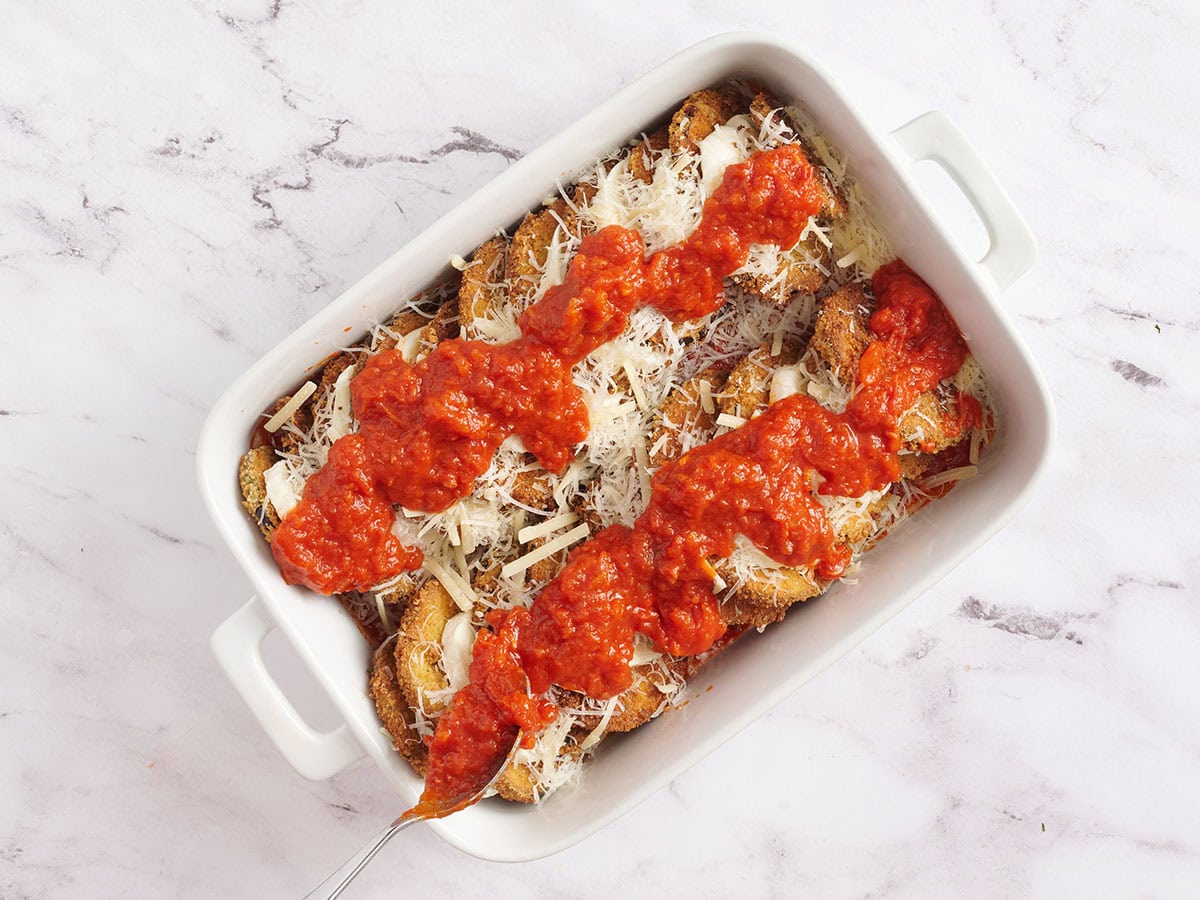 Tomato sauce over a layer of eggplant and cheese slices in white baking dish.