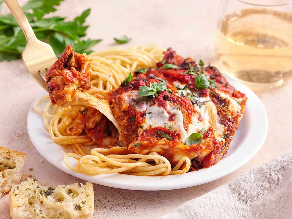 Cheese pull from a baked slice of Eggplant Parmesan served in a white dish served of spaghetti.