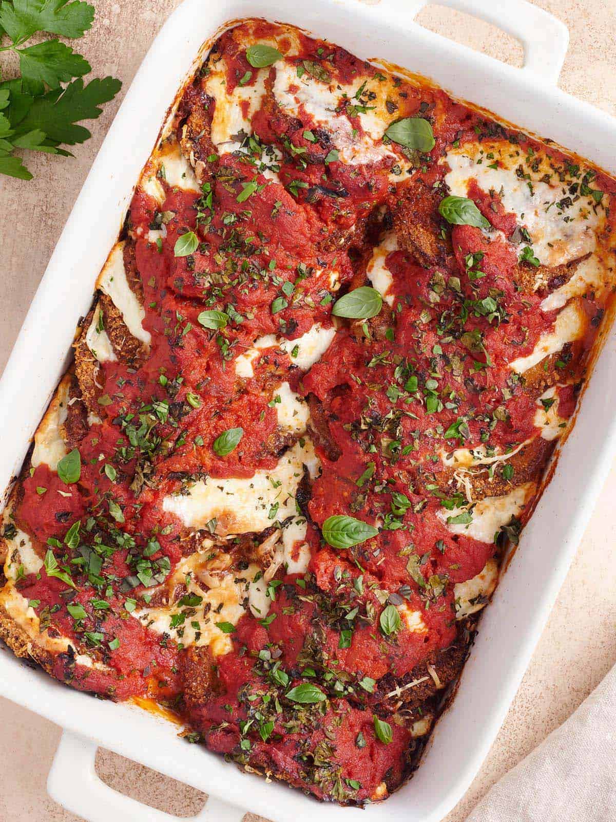 Golden-brown baked Eggplant Parmesan layered with marinara and melted cheese, garnished with fresh herbs in a white baking dish.