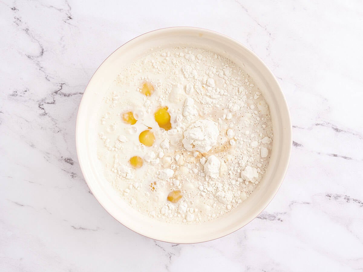 A mixing bowl with eggs, milk, flour, sugar, and salt before mixing, sitting on a white marble surface.
