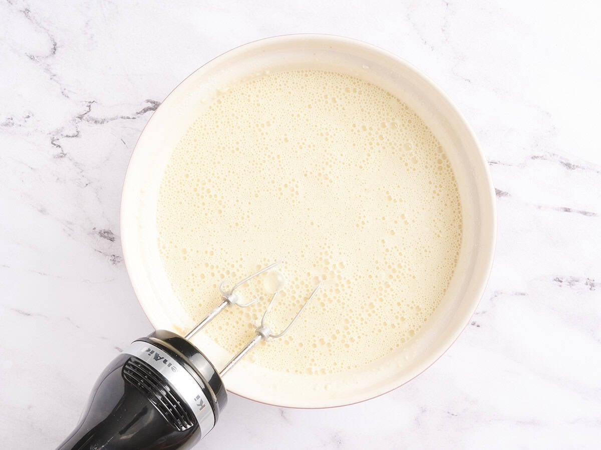 A mixing bowl of smooth crepe batter after whisking with a hand mixer, resting on a white marble surface.