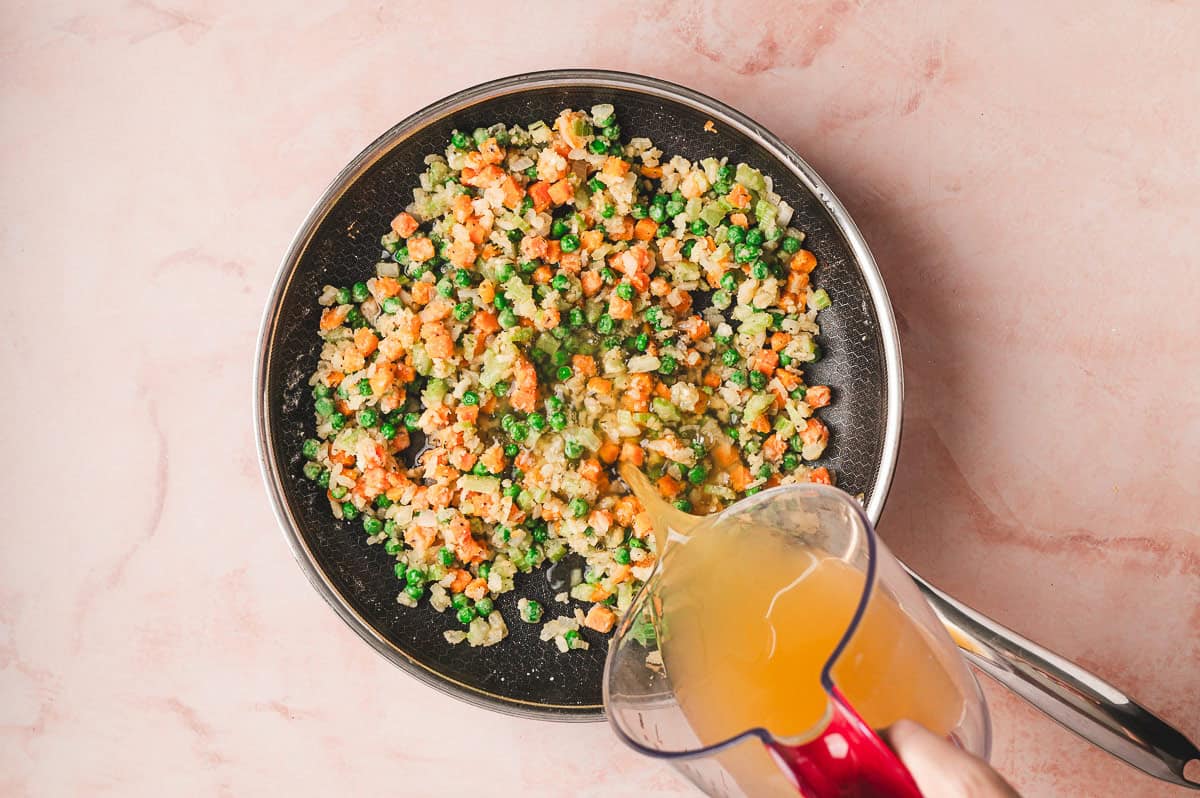 Chicken broth and milk being stirred into the vegetable mixture in the skillet to create a creamy base.