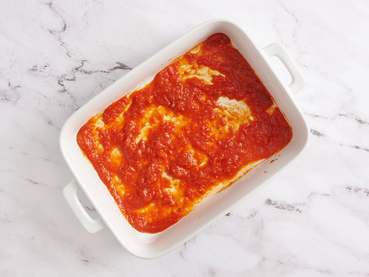 Tomato sauce spread on the bottom of a white ceramic baking dish as the first layer for Eggplant Parmesan.