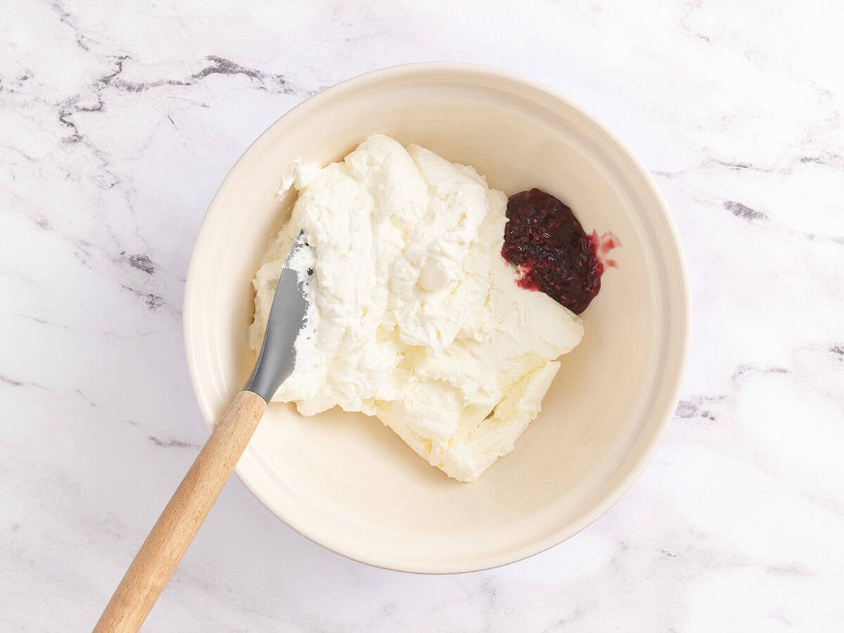 A mixing bowl with softened cream cheese and a spoonful of raspberry jam before blending.