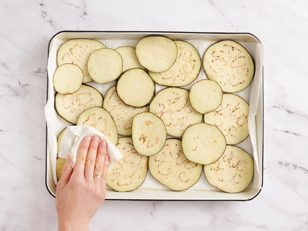 Hand pressing a paper towel on salted eggplant slices to remove moisture.