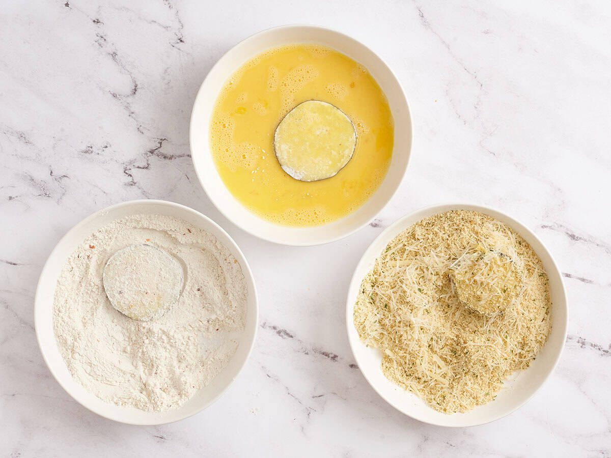 Eggplant slices dipping into the breadcrumb mixture with dredging bowls in the background.