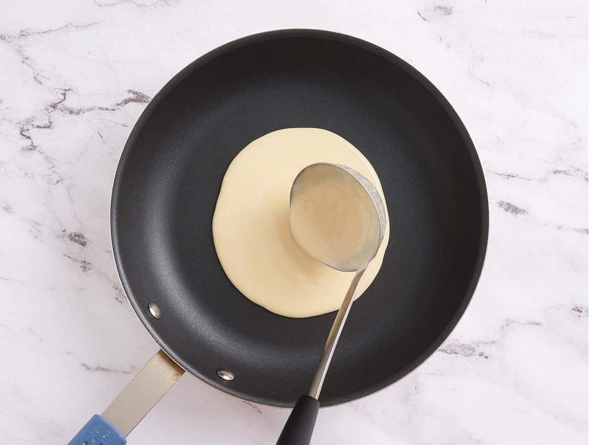 A ladle pouring crepe batter into the center of a non-stick skillet over medium heat.