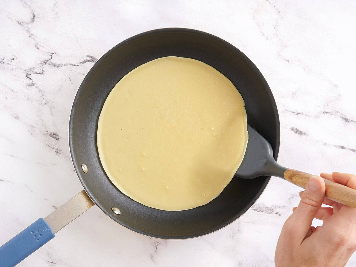 A crepe cooking in a non-stick skillet being flipped with a spatula.