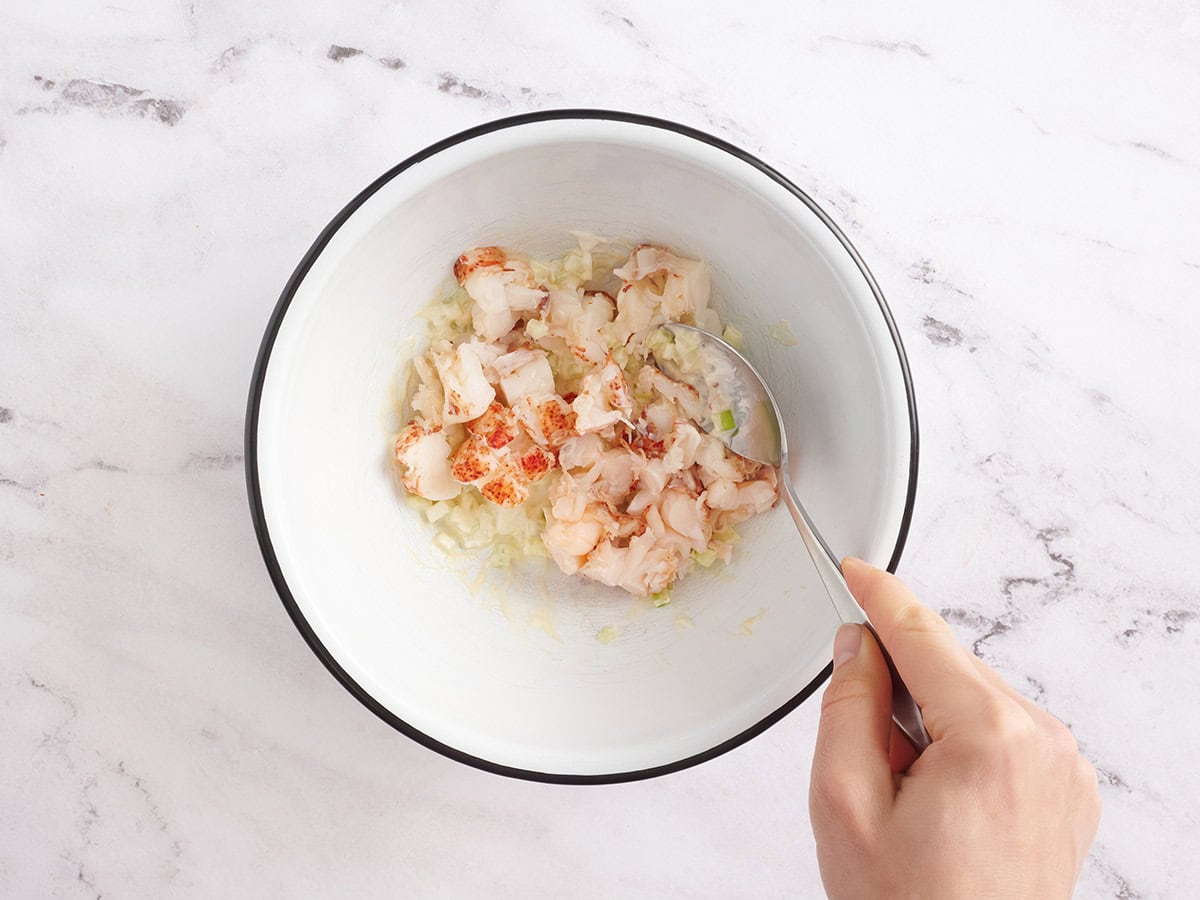 Lobster meat being mixed with mayonnaise and diced celery in a large bowl.
