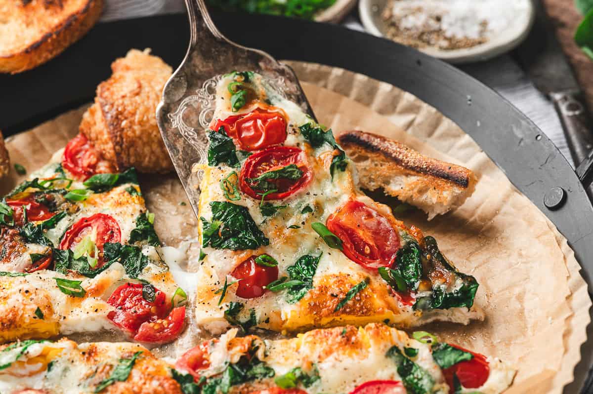 Close-up of a frittata slice being lifted with a serving spatula, showing gooey cheese, tomatoes, and spinach.