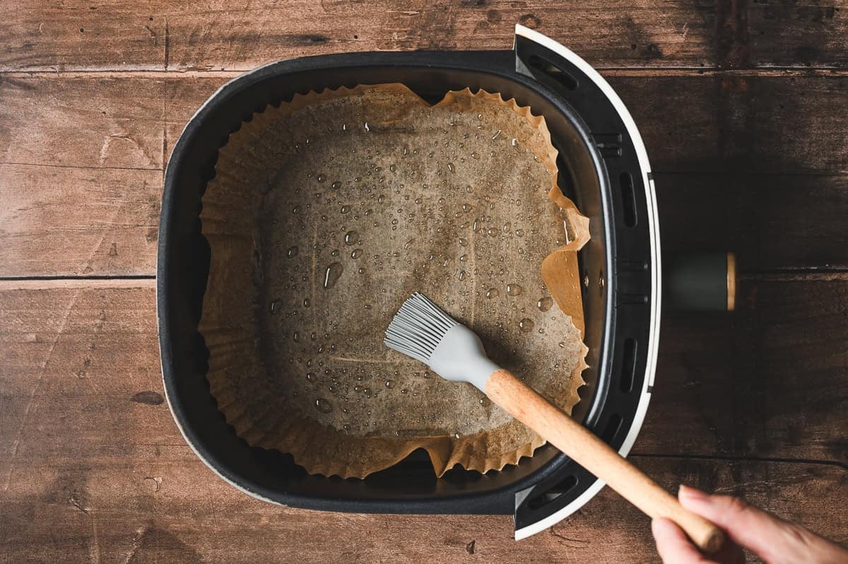 Air fryer basket lined with parchment paper being brushed with olive oil.