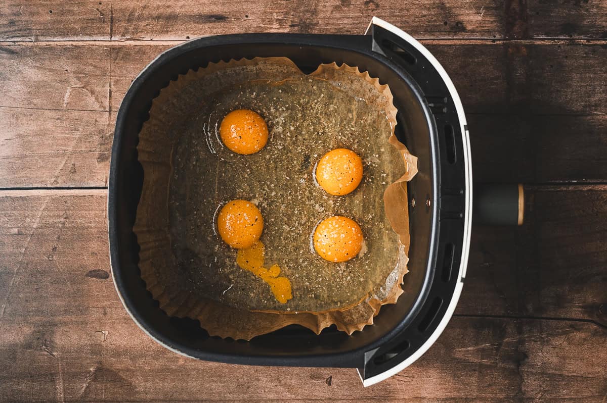 Four raw eggs cracked into the air fryer basket on top of the parchment liner, seasoned with salt and pepper.