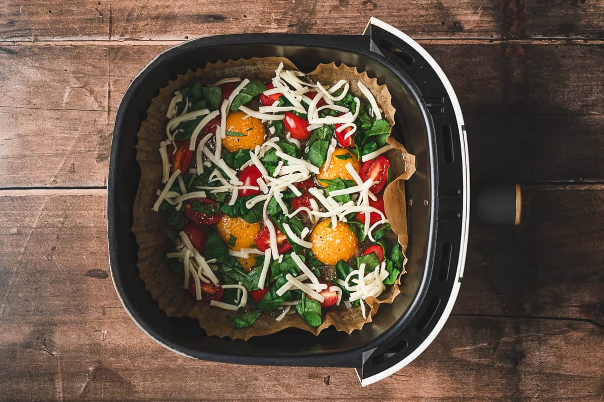Eggs in the air fryer basket topped with halved cherry tomatoes, chopped spinach, and shredded mozzarella.