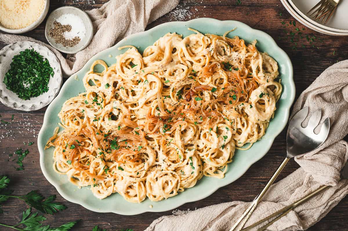 Large serving platter filled with caramelized onion pasta, topped with extra onions and parsley, with small bowls of Parmesan, parsley, and seasonings on the side.