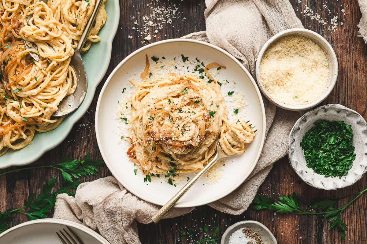 A white plate of creamy caramelized onion pasta twirled with a fork, topped with Parmesan and parsley, with extra grated cheese and herbs in small bowls nearby.