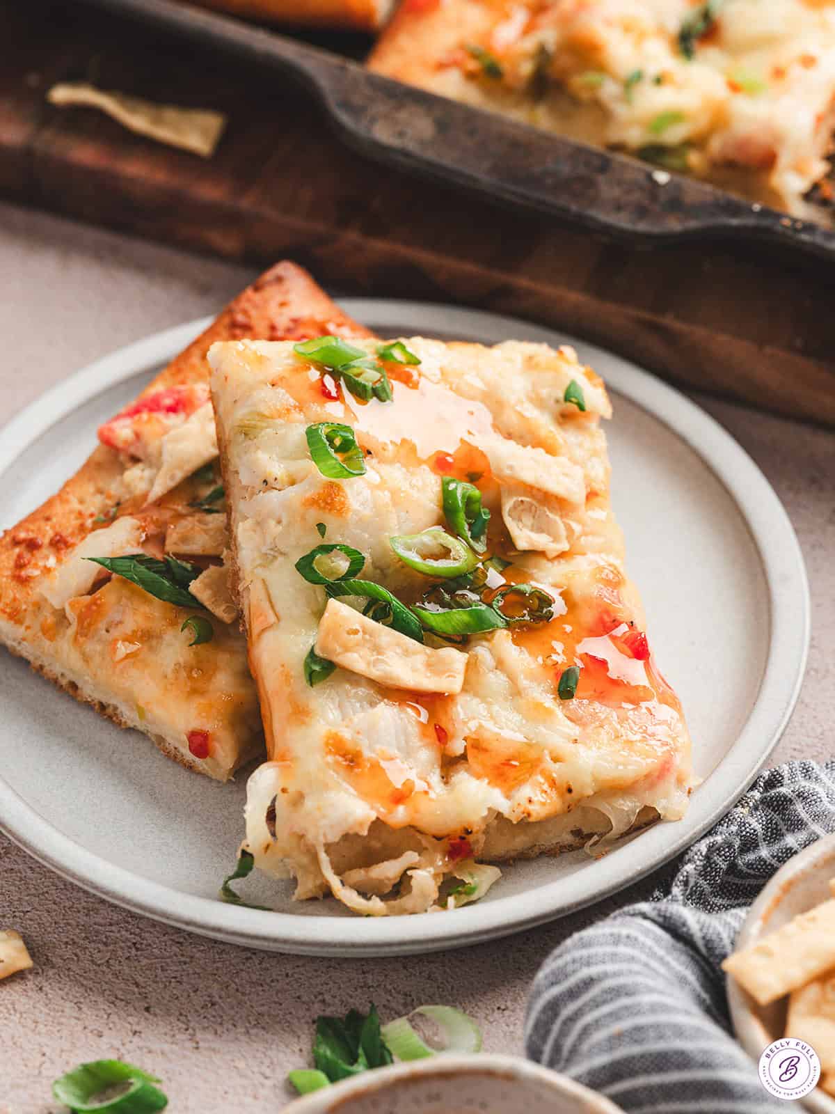 Two slices of crab rangoon pizza on a white plate, garnished with green onions and sweet chili sauce drizzle.