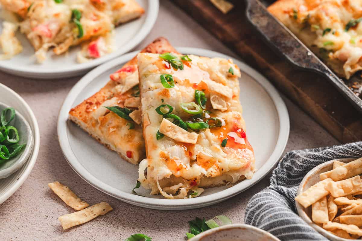 Two slices of crab rangoon pizza on a white plate, garnished with green onions and sweet chili sauce drizzle.