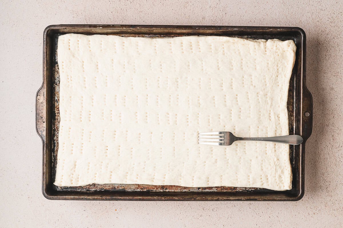 Rectangle of pizza dough pressed into a sheet pan and docked with a fork before baking.